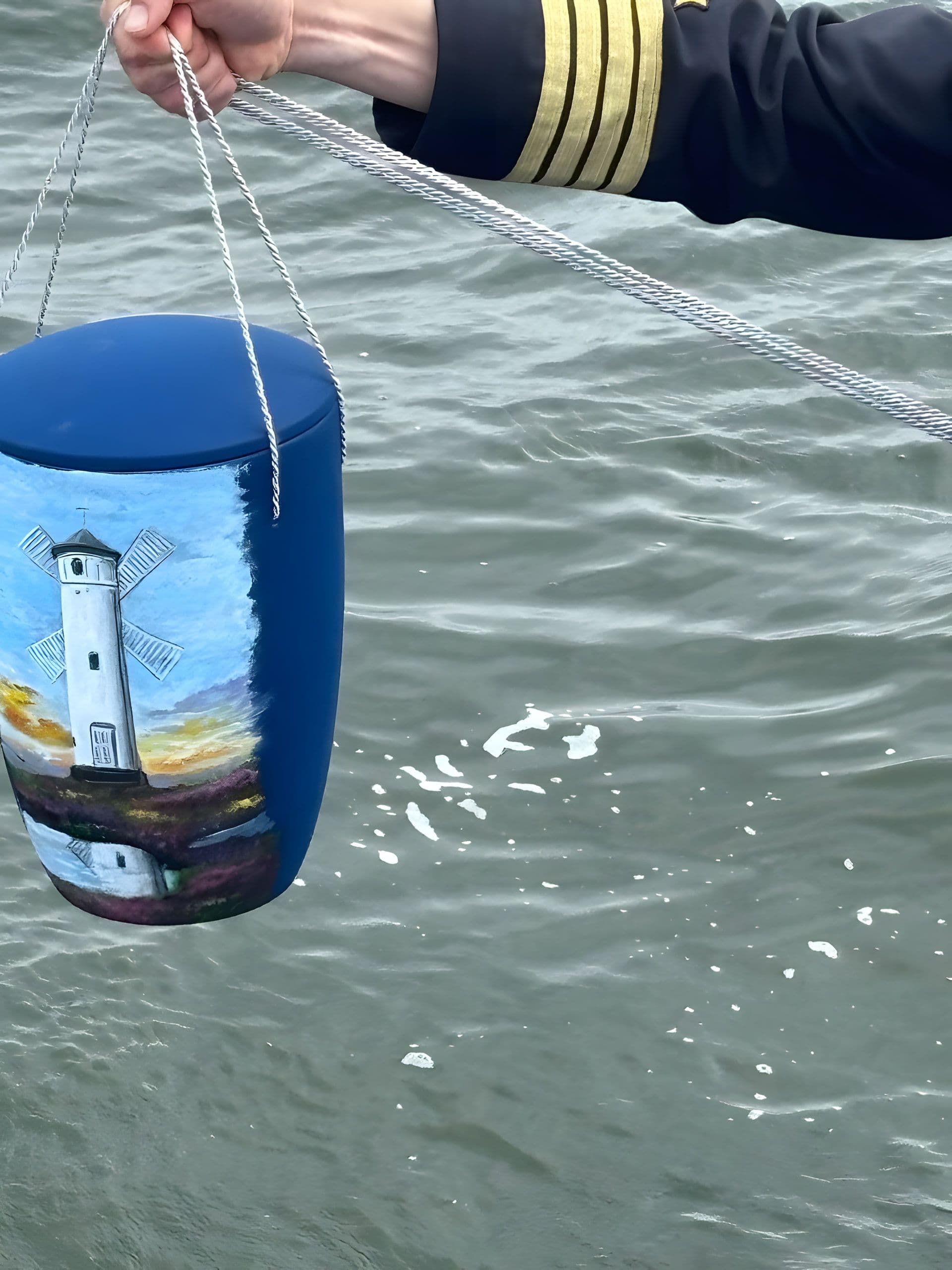 Blau bemalte Seeurne mit Leuchtturm und Windmühle, an Leinen über dem Wasser